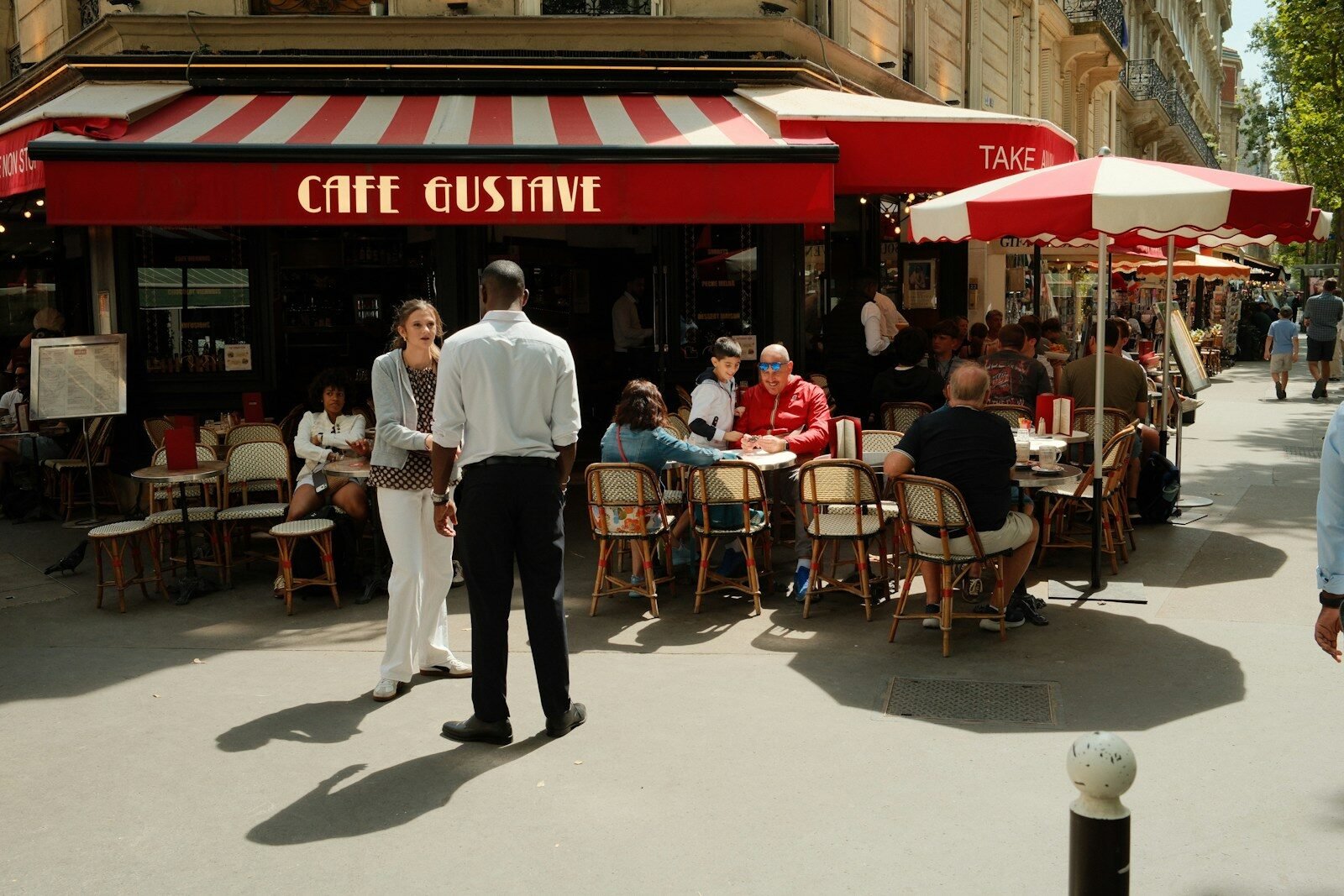 People sitting at tables outside Cafe Gustave on a sunny sidewalk in Paris