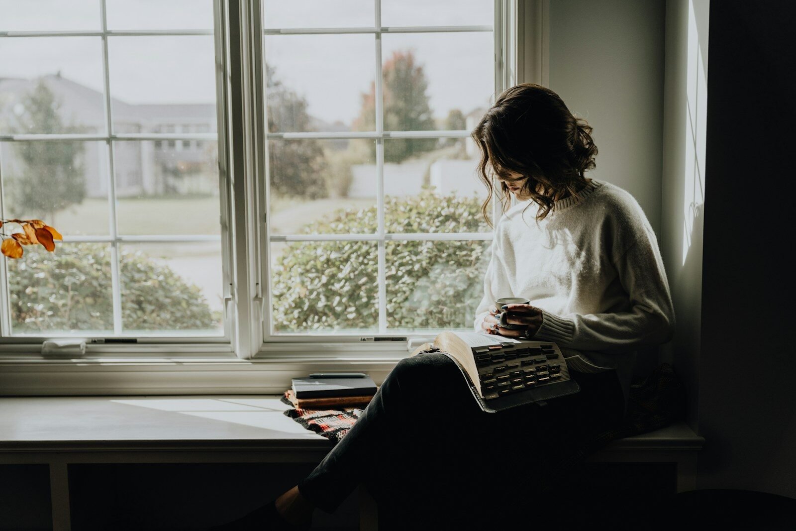 A woman in a white sweater holding a mug while reading a book